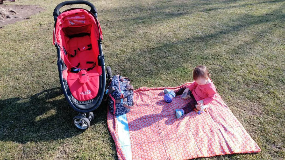 Baby girl sits on picnic blanket next to jogging stroller.