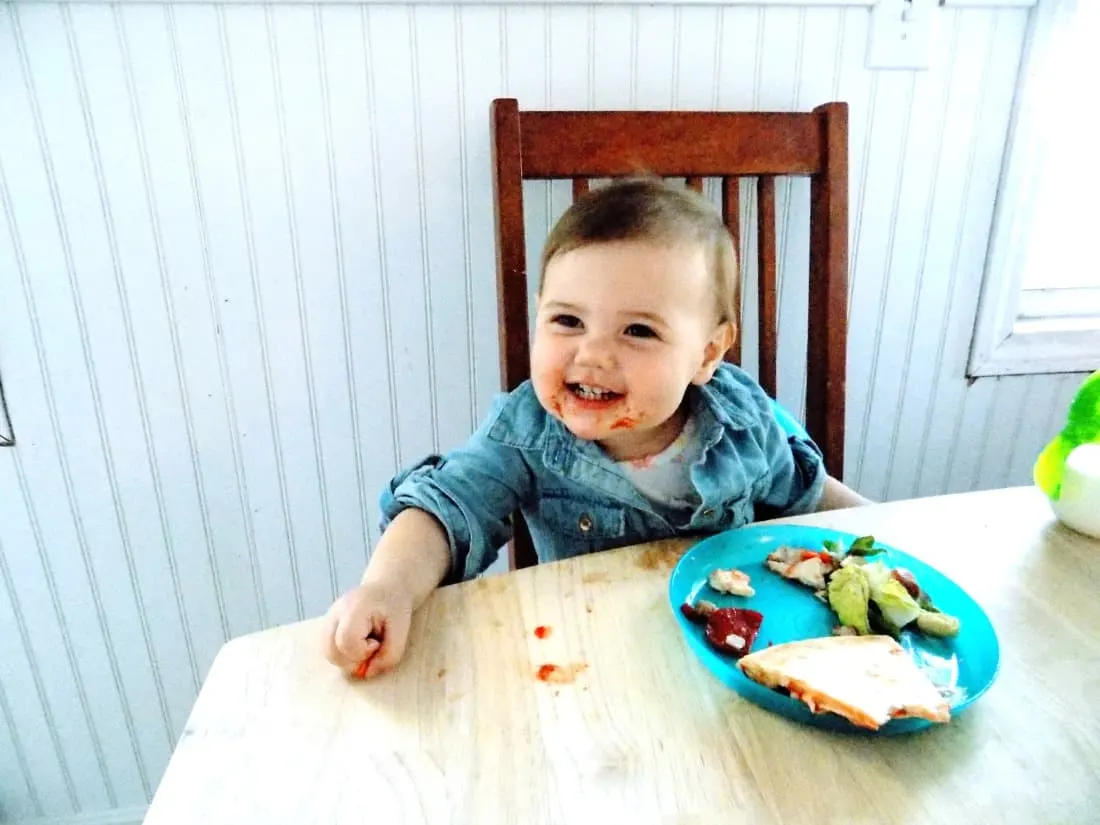 Toddler girl eats pizza at dining table.