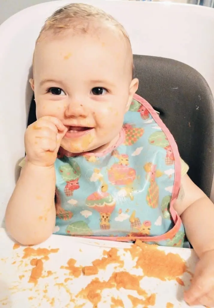 Baby girl messily eats food in high chair.