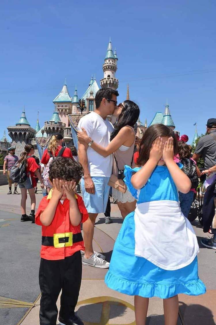 Parents kiss in front of Disney castle while children cover their eyes.