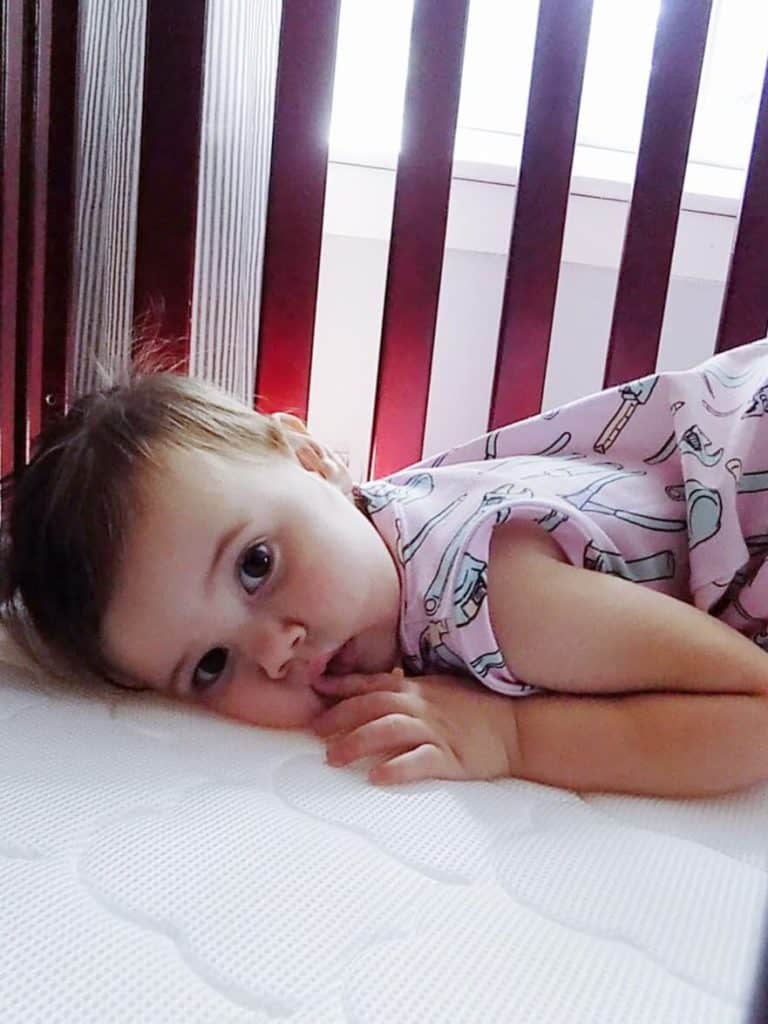 Toddler girl lays in crib and stares at camera.