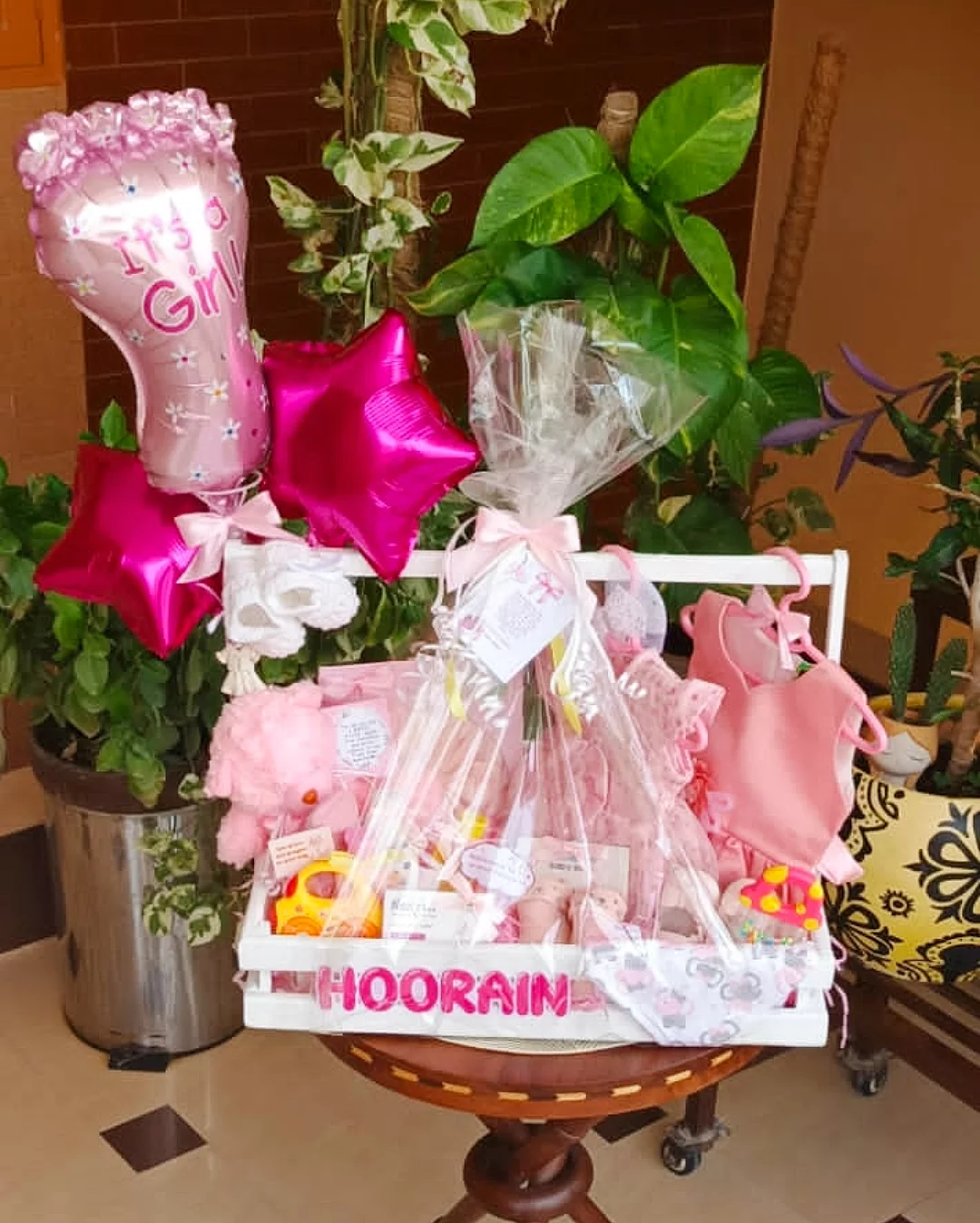 A baby shower gift in a wooden crate with a rod with hanging cloths and pink balloons.