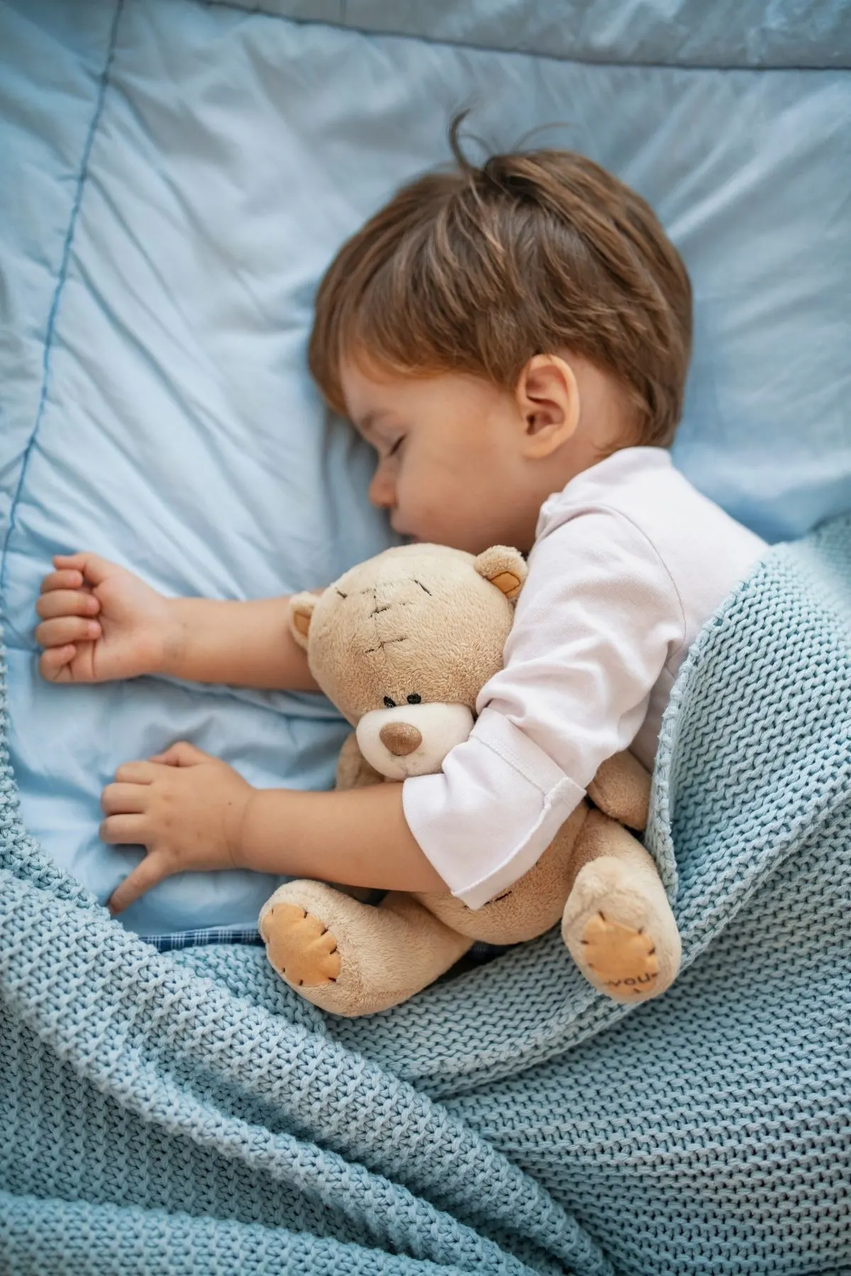 Toddler boy sleeping while snuggling teddy bear with teal blanket.
