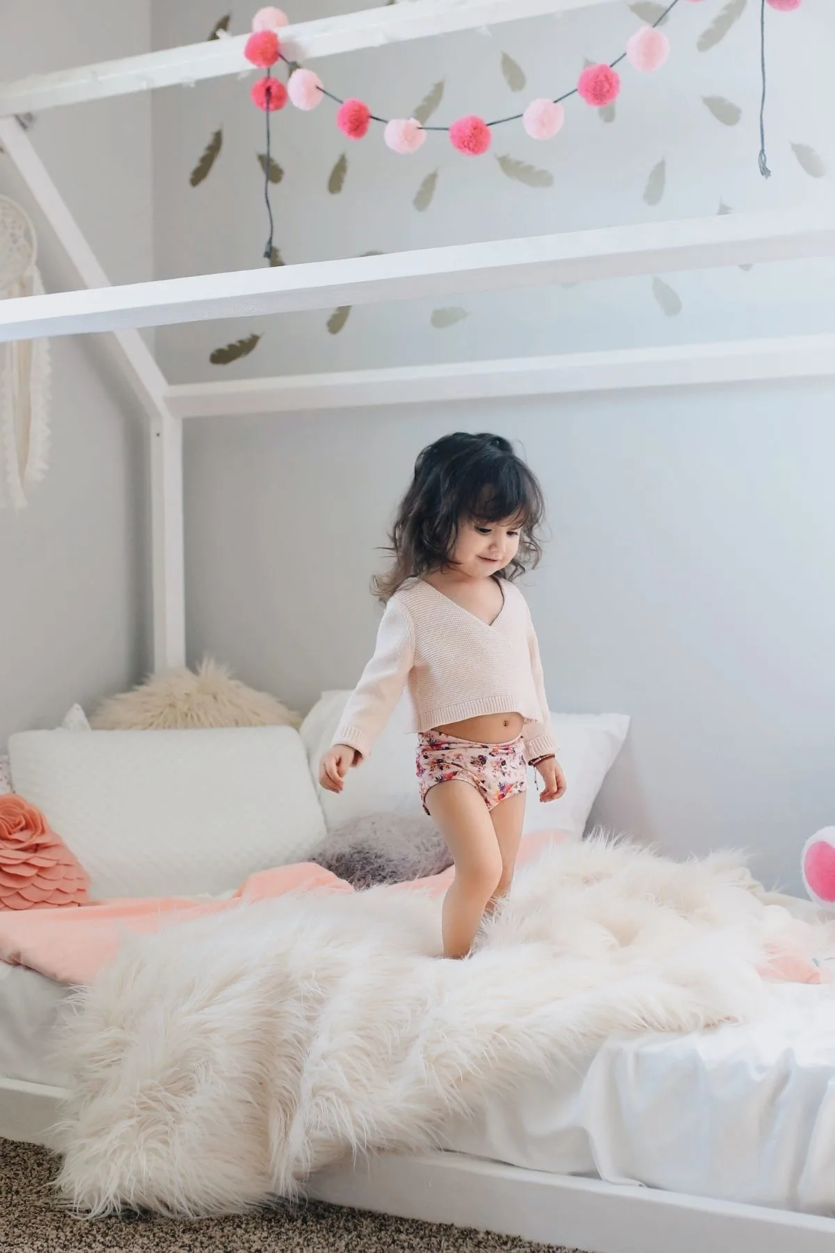 Toddler girl stands on her bed with pink and white sheets and decorations.