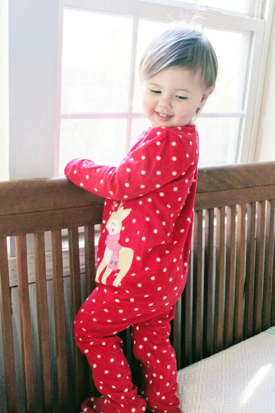 Toddler girl in Christmas pajamas stands in crib.