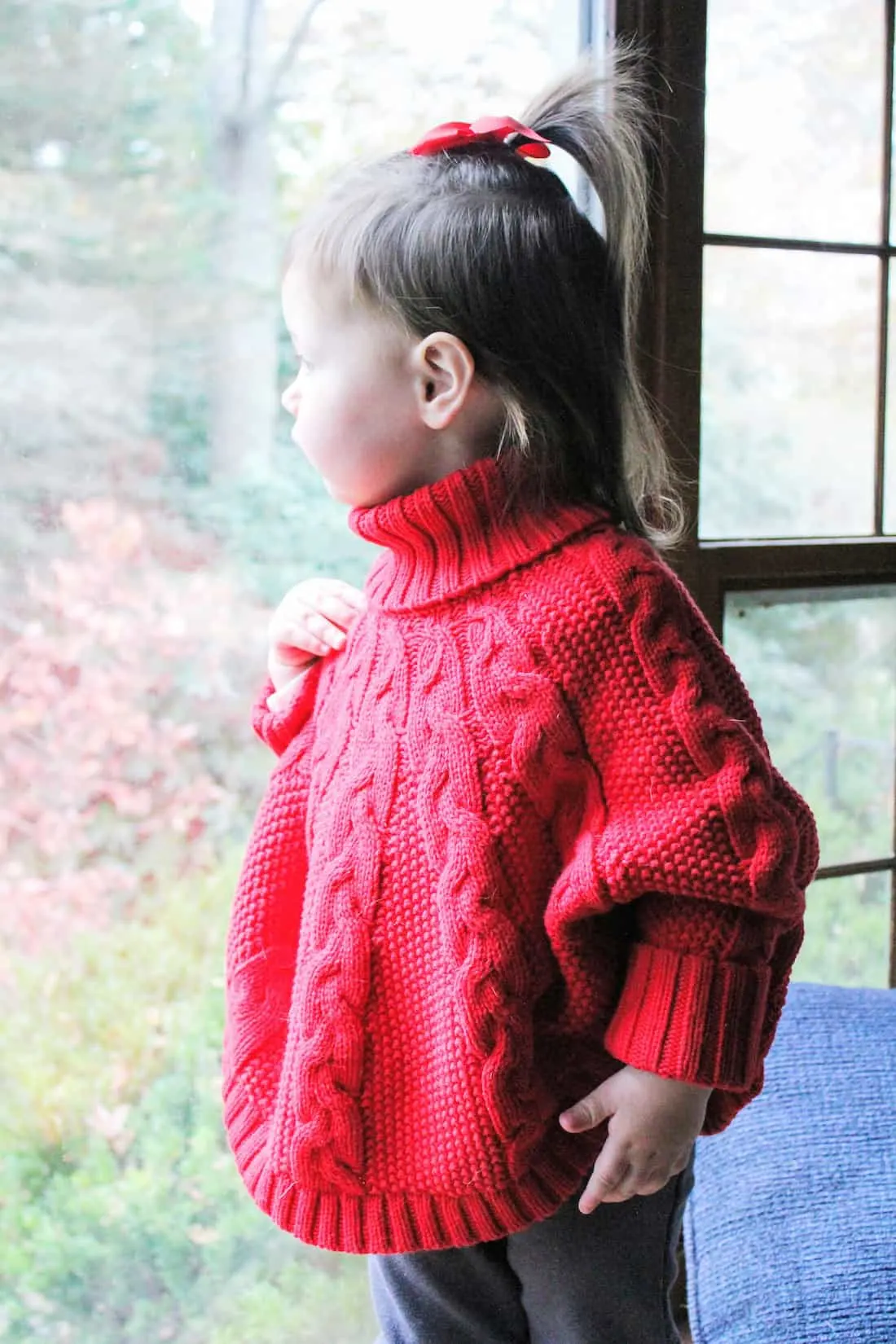Toddler girl in red holiday sweater looks through window.