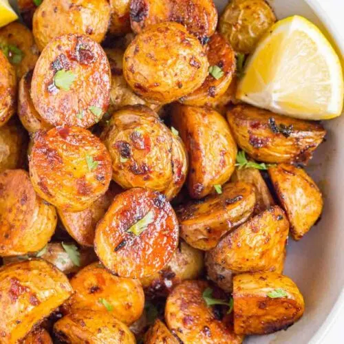 Harissa roasted potato wedges in a white bowl with green garnish and a lemon wedge.