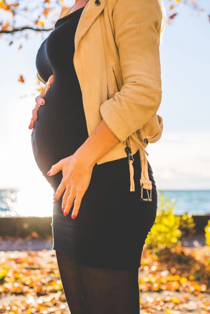 A pregnant woman in a black shirt and pants, with a tan jacket outside during fall.