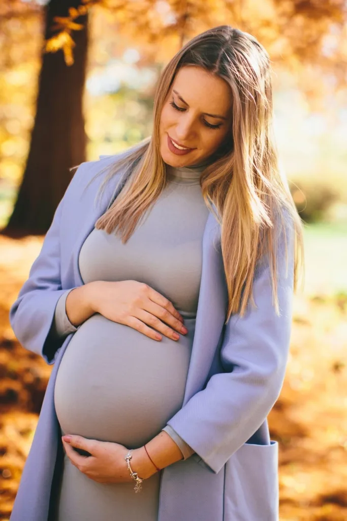 A pregnant woman in a gray dress and blue coat outside during fall.