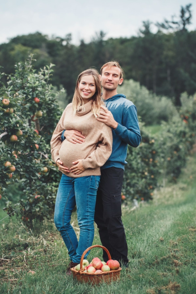 A pregnant woman and her partner pose in an apple orchard.