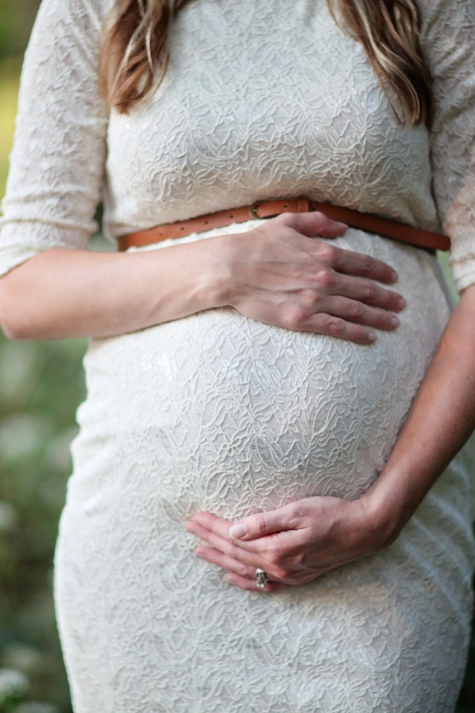 A pregnant woman in a lacy white dress with a brown belt outside.