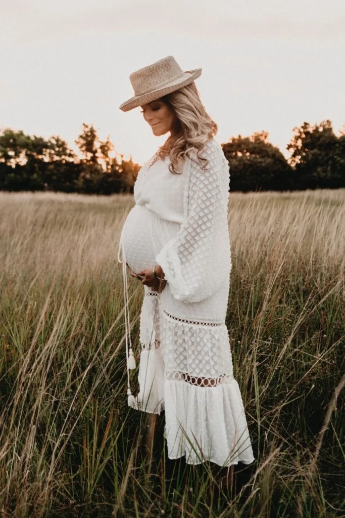 A pregnant woman in a flowy white dress and a hat with a brim outside.