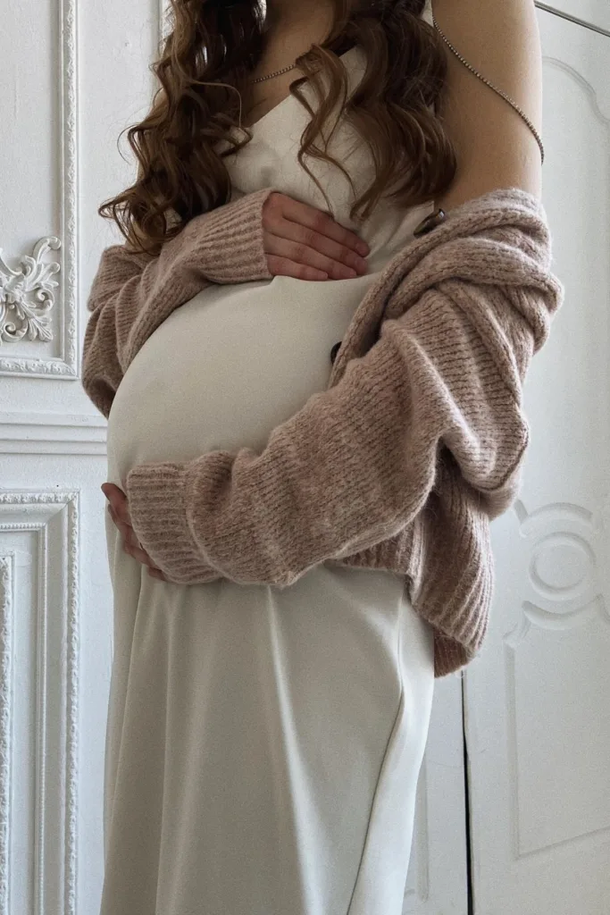 A pregnant woman in a tan cotton dress and a dusty pink off-the-shoulder sweater.