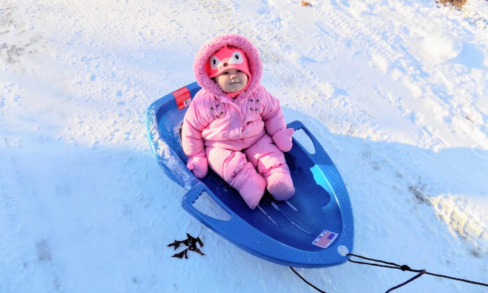 Baby girl sits on sled in snow.