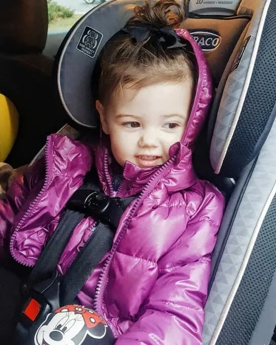 Toddler girl in puffer coat strapped into car seat.