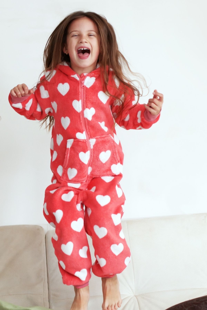 A girl jumps on a couch in red onesie pajamas with white hearts.