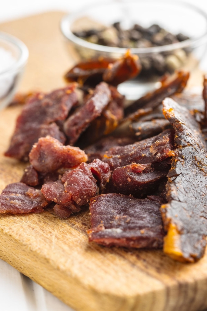Slices of beef jerky on a wooden cutting board next to a bowl of peppercorns.