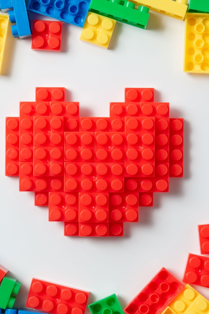 Red Legos in the shape of a heart with other Lego bricks on a white table.