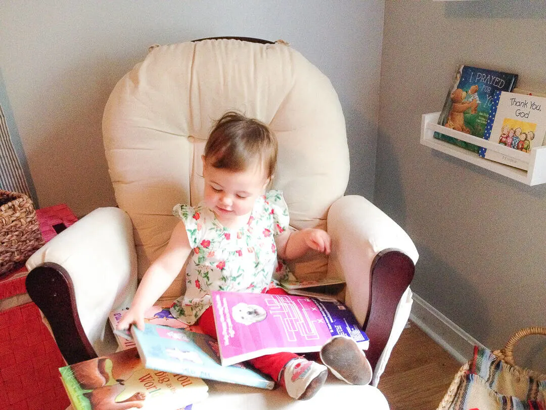 Little girl sits on rocking chair glider with a lap filled with books.