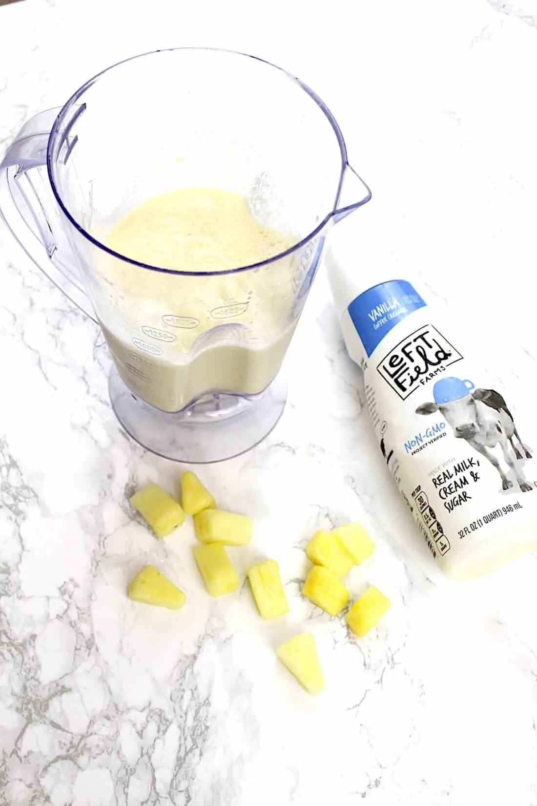 Smoothie in blender next to pineapple chunks and milk bottle.