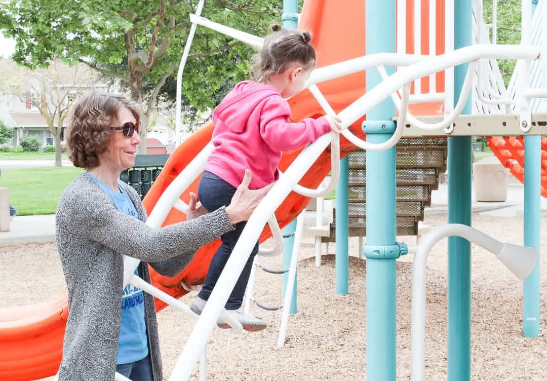Woman helps toddler girl onto playground latter.
