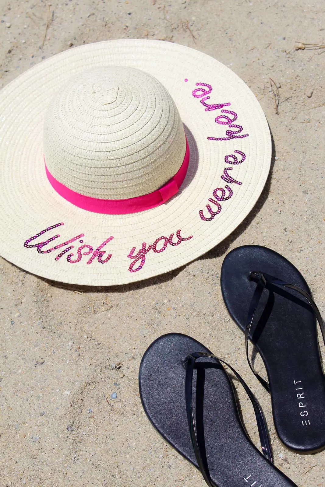 Wish you were here summer hat lays on beach next to black flip flops.