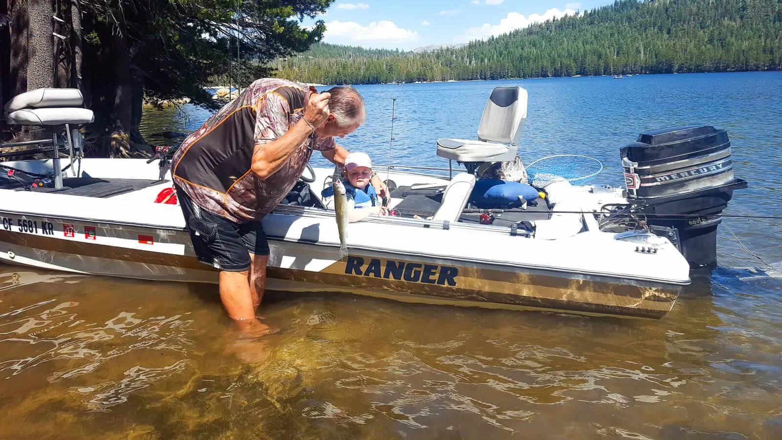 Man holds fish on fishing line next to small boat with toddler girl inside.