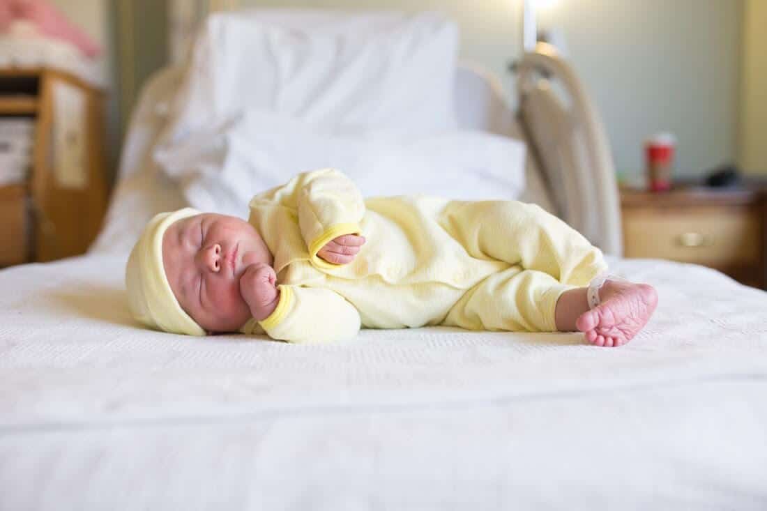 Newborn in yellow onesie and hat sleeps on bed.