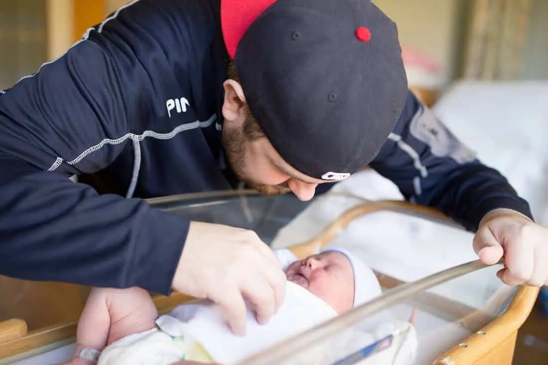 Man looks over newborn baby in hospital bassinet.
