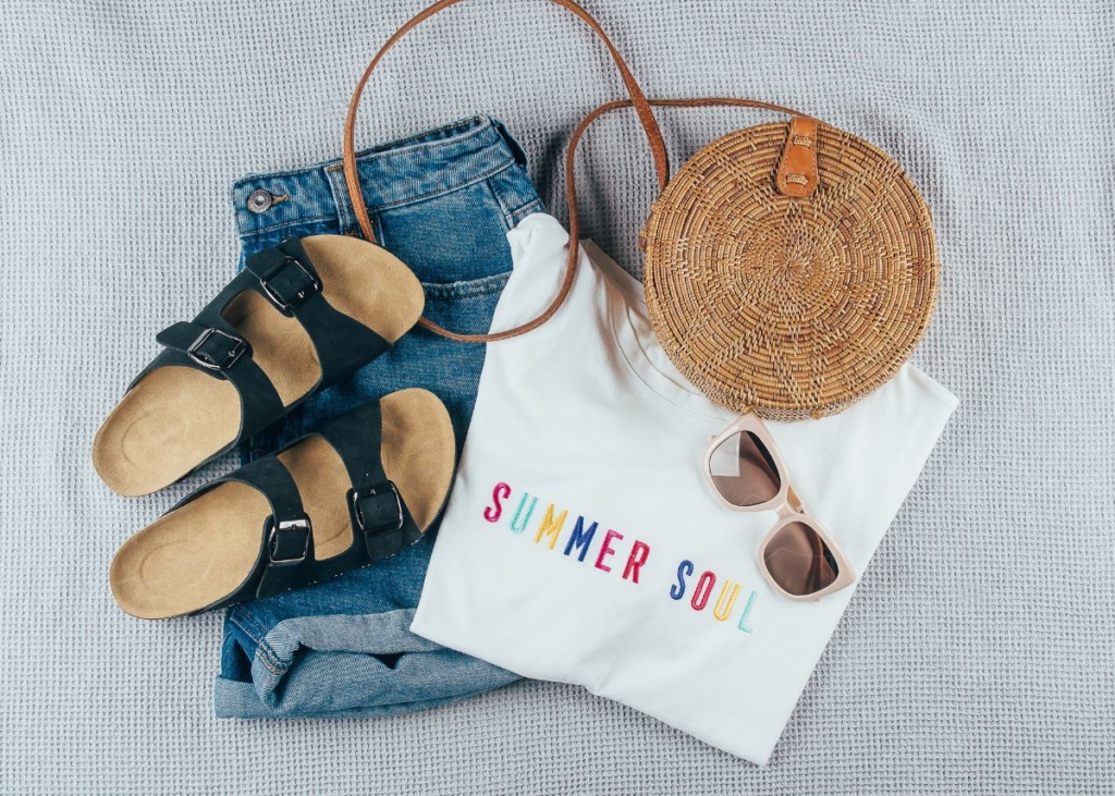 Flat lay outfit with jean shorts, black sandals, and a white shirt with colorful wording.