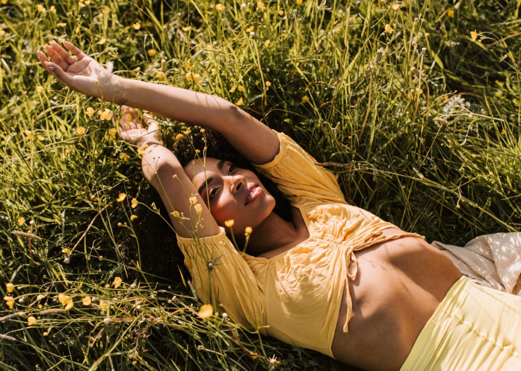 A woman in a yellow summer outfit laying in a field of grass and wildflowers.