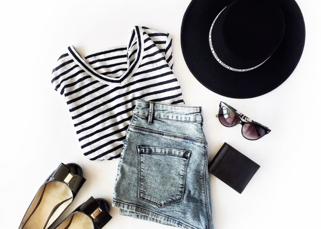Flat lay outfit with a black and white striped shirt, jean shorts, black flats, and a black hat.
