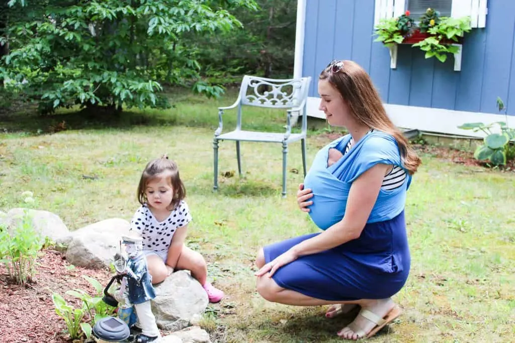 Mother plays with daughter outdoors and holds baby in carrier.