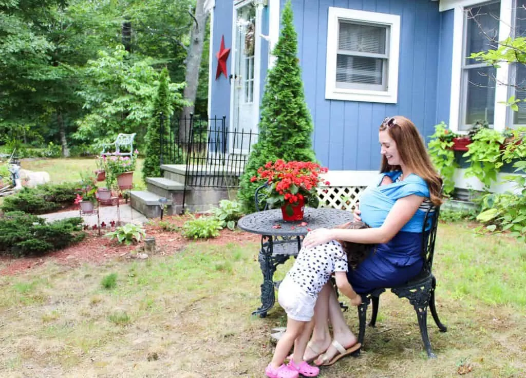 Mother holds baby in carrier sling while parenting toddler in yard.