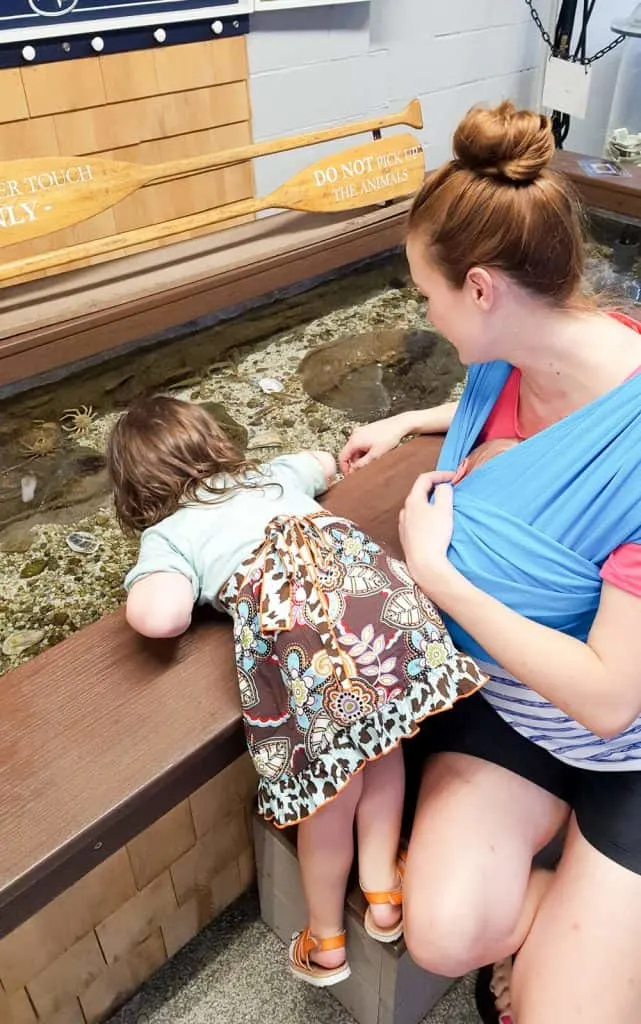 Mother uses sling to carry new baby while engaging with toddler at zoo.