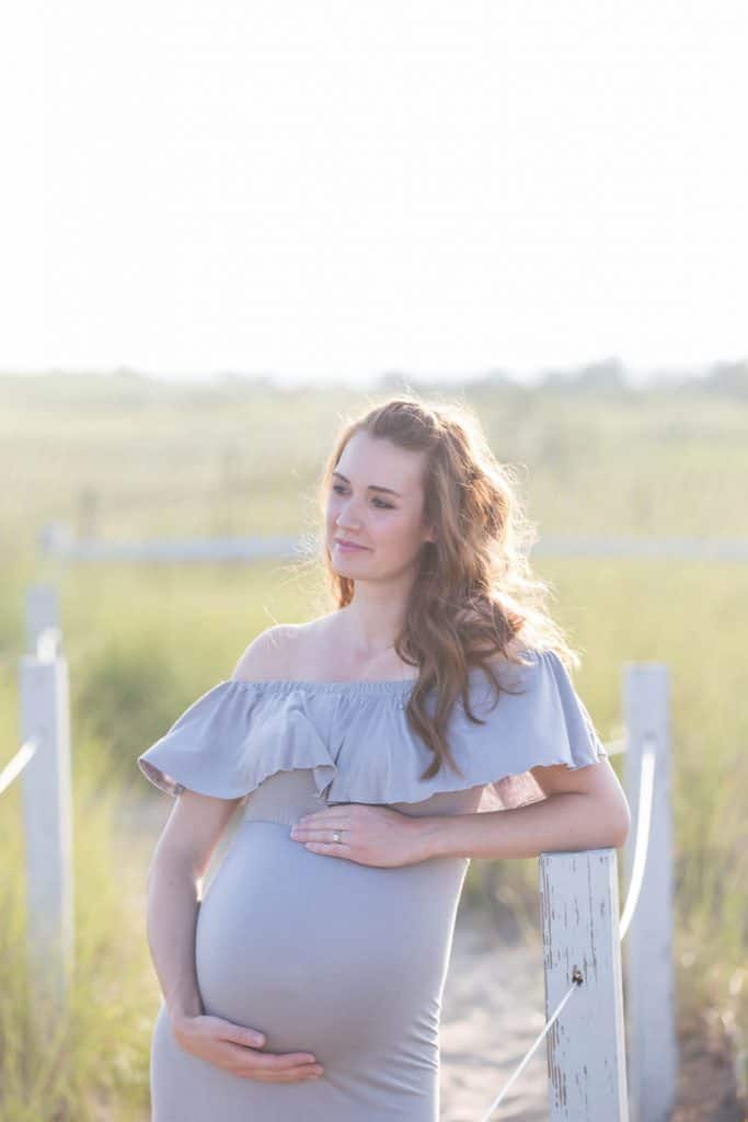 Woman poses next to tall beach grass for maternity pictures.