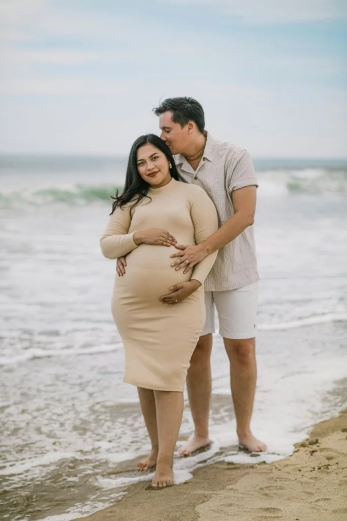 A couple, including a pregnant woman in a tan dress pose the ocean's edge on a beach.