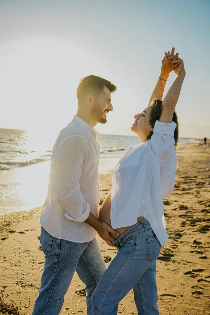 A couple, including a pregnant woman in white shirts and jeans posing on the beach.