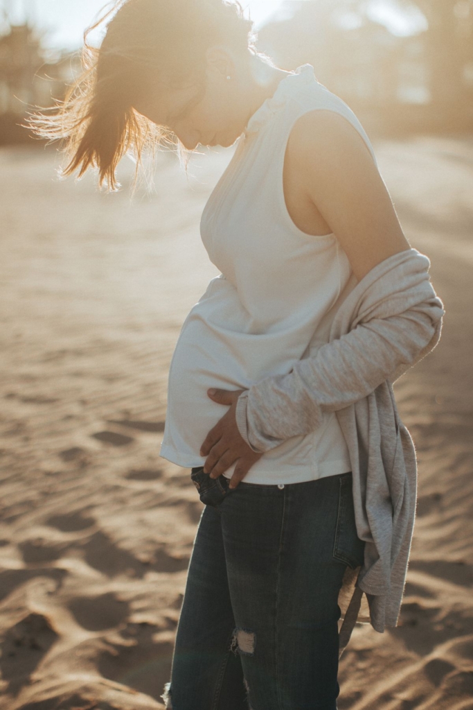 A pregnant woman in a white tank and jeans posing on a beach.
