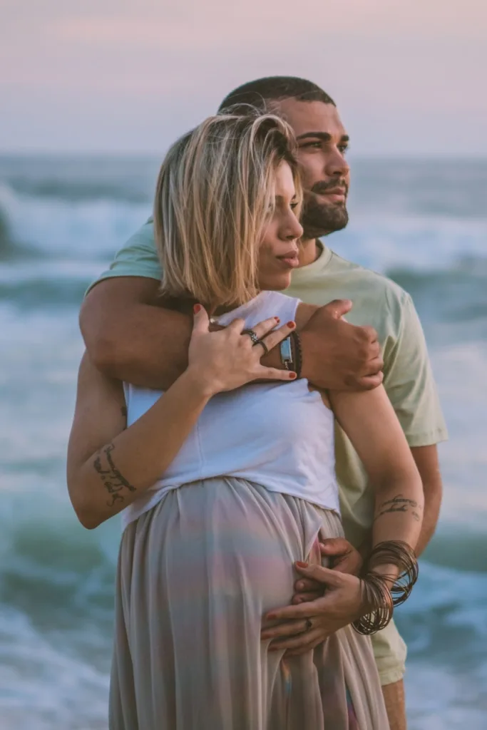 A couple, including a pregnant woman embrace on the beach.