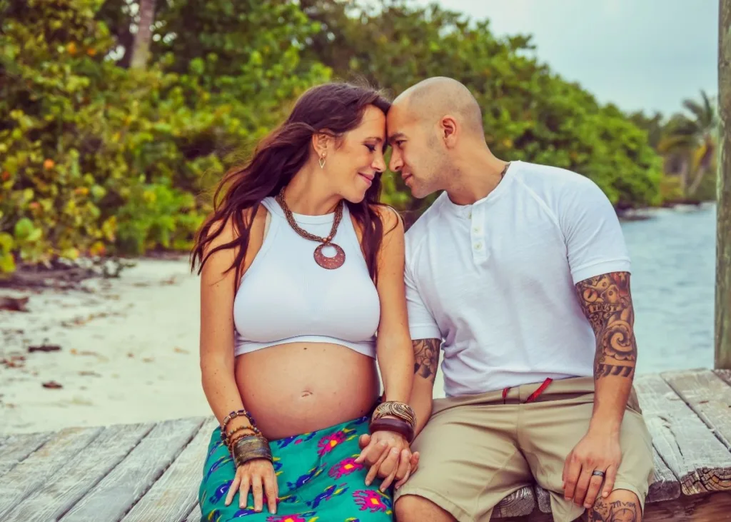 A couple sits on a dock at the beach, posing for maternity photos.