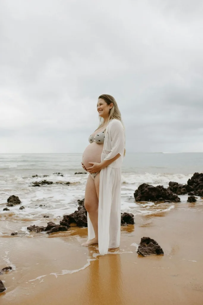 A pregnant woman in an olive green bikini and sheer white cover up posing on the beach.