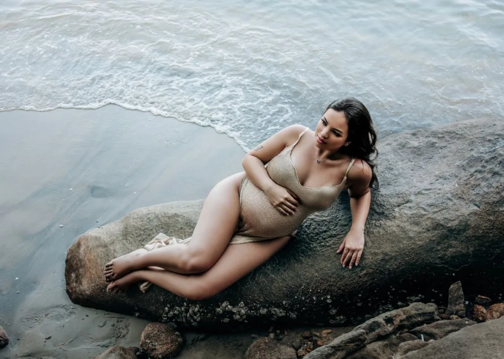 A pregnant woman in a cream bikini poses on rocks next to the ocean on the beach.