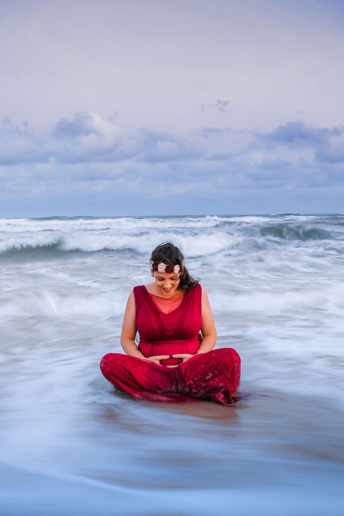A pregnant woman in a red dress, sitting crossed leg on the beach with the water washing over her.
