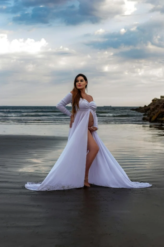 A pregnant woman in a long flower white gown posing in the ocean surf.