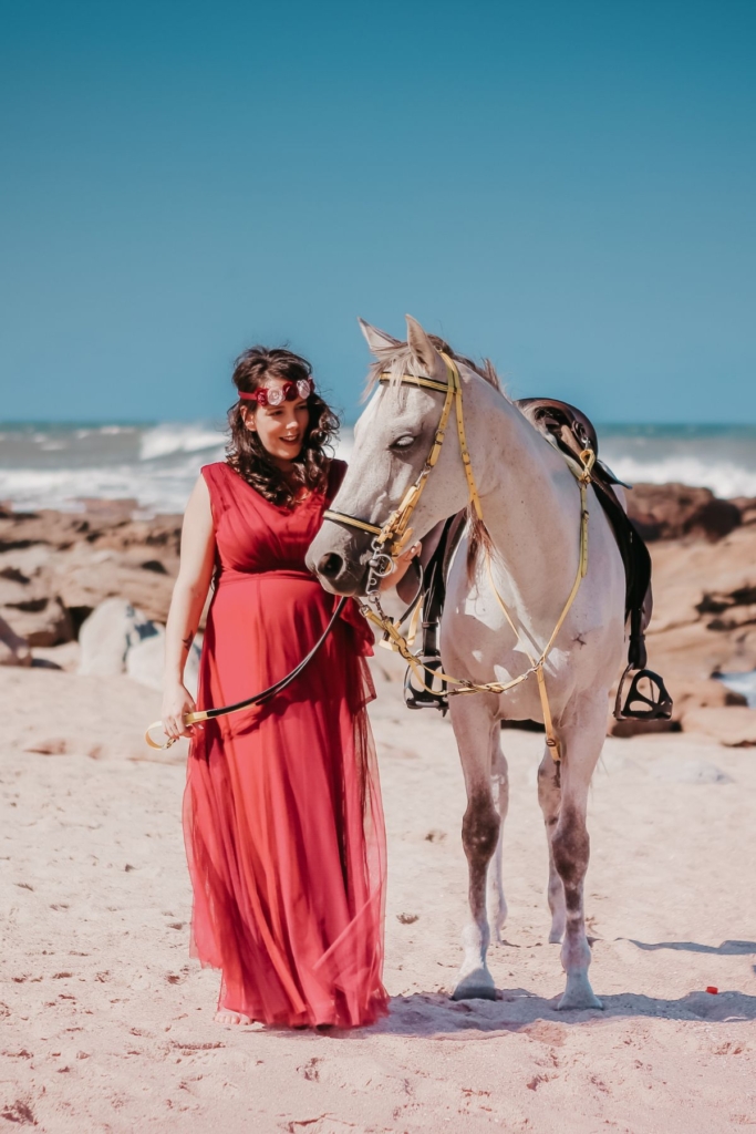 A pregnant woman in a red dress with a white horse on a beach.
