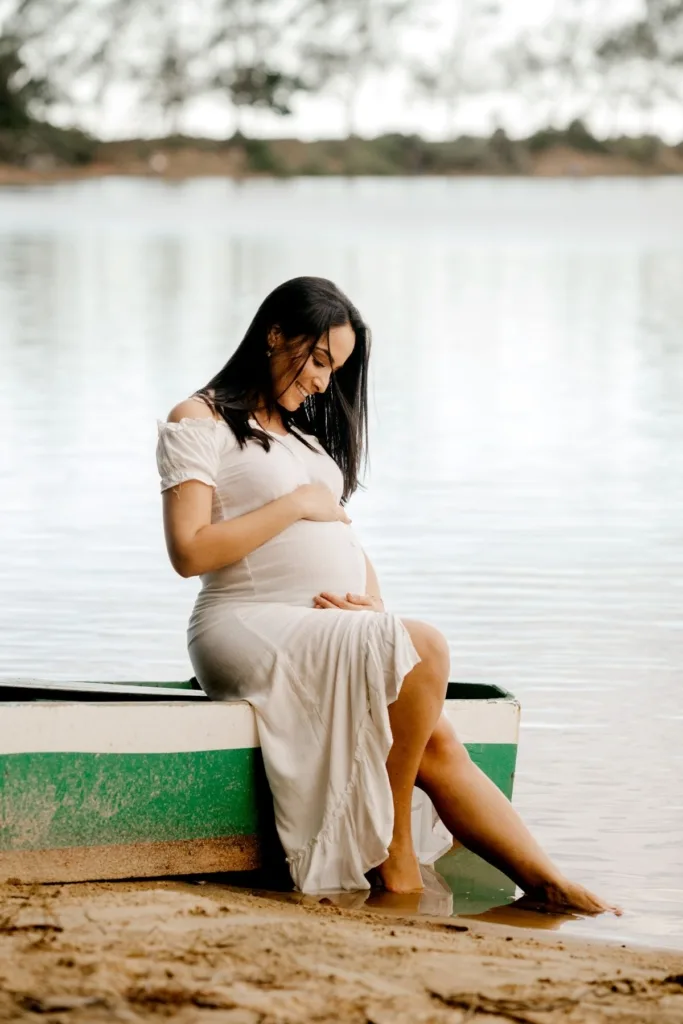A pregnant woman in a long cream dressing sitting on the edge of a canoe by the waters edge.