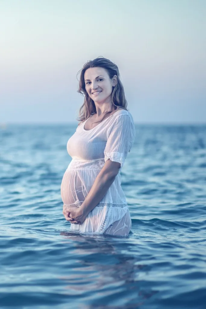 A pregnant woman in a white dress waist deep in the water.