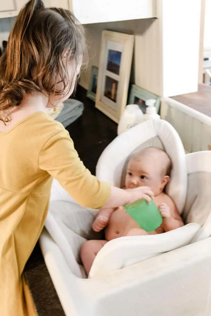 Toddler girl pours water over baby in bath.