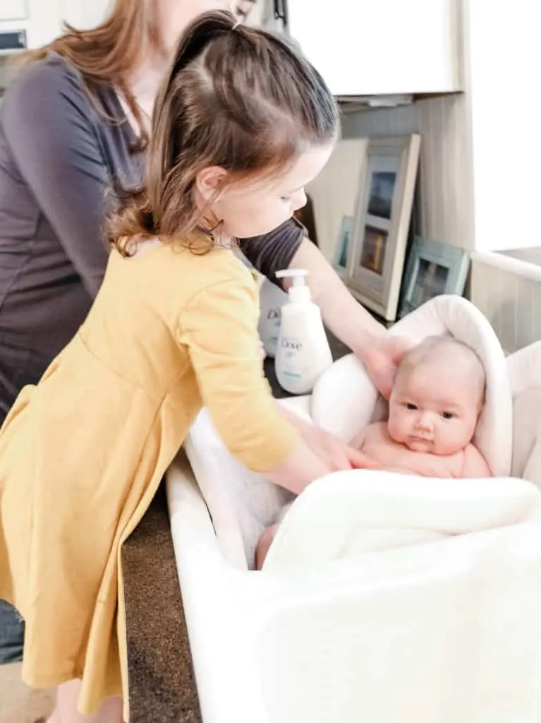 Toddler girl helps mother bathe baby in sink.